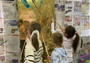 Trzy dziewczynki malują farbami po lustrze umieszczonym na ścianie z gazet