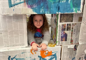 Dziewczynka patrzy przez okno utworzone z pudełek owiniętych gazetami