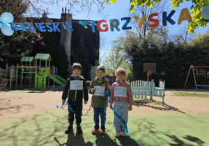 Trójka dzieci - reprezentanci grupy - stoi na tle dekoracji „Niebieskie Igrzyska”.