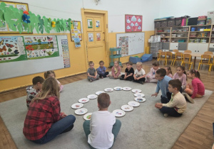 Dzieci i nauczycielka siedzą w kręgu na dywanie, wewnątrz którego ułożone są papierowe talerzyki z obrazkami.