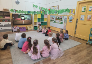 Przedszkolaki siedzą na dywanie w sali i oglądają filmik o autyźmie na dużym ekranie multimedialnym.