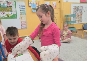 Dziewczynka w grubych rękawicach kuchennych próbuje rysować kredką na kartce leżącej na krześle.