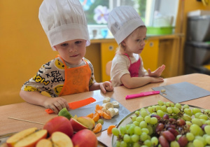 Chłopiec kroi pomarańczowym nożem banana, a dziewczynka w różowym fartuszku patrzy na owoce przed nią.