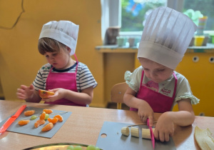 Dwie dziewczynki w różowych fartuszkach i kucharskich czapkach skupione na swoich zadaniach; jedna kroi różowym nożem banana, a druga nabija cząstki mandarynki na patyczek.