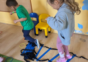 Dwoje dzieci ćwiczy na podłodze, biegając i skacząc przez niebieską drabinkę koordynacyjną.
