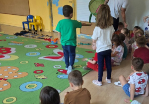 Grupa dzieci siedzi na dywanie w sali przedszkolnej, a trener przybija z nimi „piątki”.