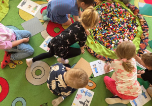 Dzieci siedzą na dywanie wokół dużego worka z klockami LEGO, bawiąc się i budując.