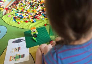 Dziewczynka siedzi na dywanie i buduje konstrukcję z klocków LEGO.
