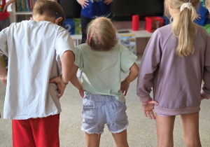 Widok od tyłu na troje przedszkolaków stojących w rozkroku z rękami opartymi na biodrach. Przed nimi stoi instruktorka Zumby w niebieskiej koszulce z napisem „Taniec to moja supermoc”.