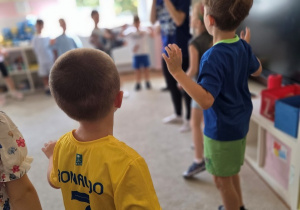 Chłopiec w żółtej koszulce sportowej z numerem 7 tańczy w sali przedszkolnej, Unosząc ręce w stronę instruktorki prowadzącej zajęcia Zumby.