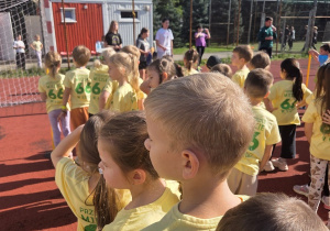 Grupa dzieci w żółtych koszulkach stoi na boisku sportowym, czekając na rozpoczęcie konkurencji.