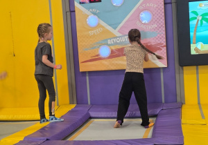 Dwie dziewczynki skaczą na małych trampolinach, próbując dotknąć świecących punktów na interaktywnej, kolorowej tablicy z wynikami i czasem na ścianie.