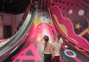 Dzieci w sali zabaw stoją pod wielką, różowo-czerwoną zjeżdżalnią w kształcie wulkanu z uchwytami do wspinaczki; jedno z nich właśnie zjeżdża z góry.