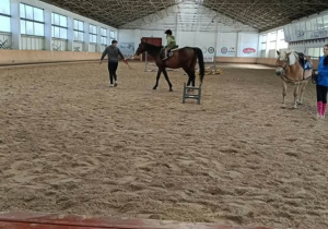 Dziecko jeździ na koniu pod opieką instruktora w dużej krytej ujeżdżalni.